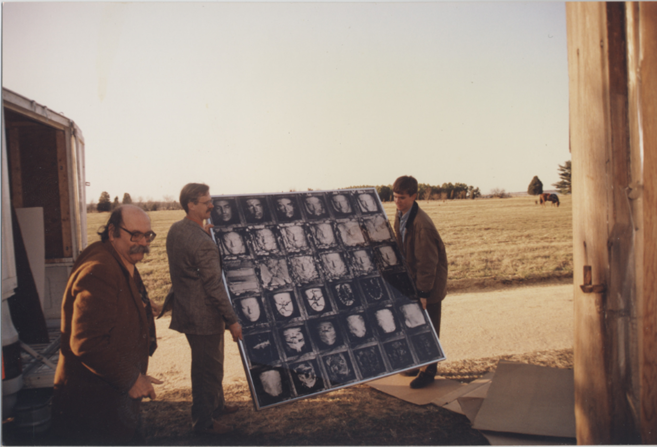 Photograph showing three men carrying a large board with multiple black-and-white photographic portraits arranged in a grid. The setting is an outdoor rural area with open fields and a clear sky, suggesting transport or installation of the portrait display.