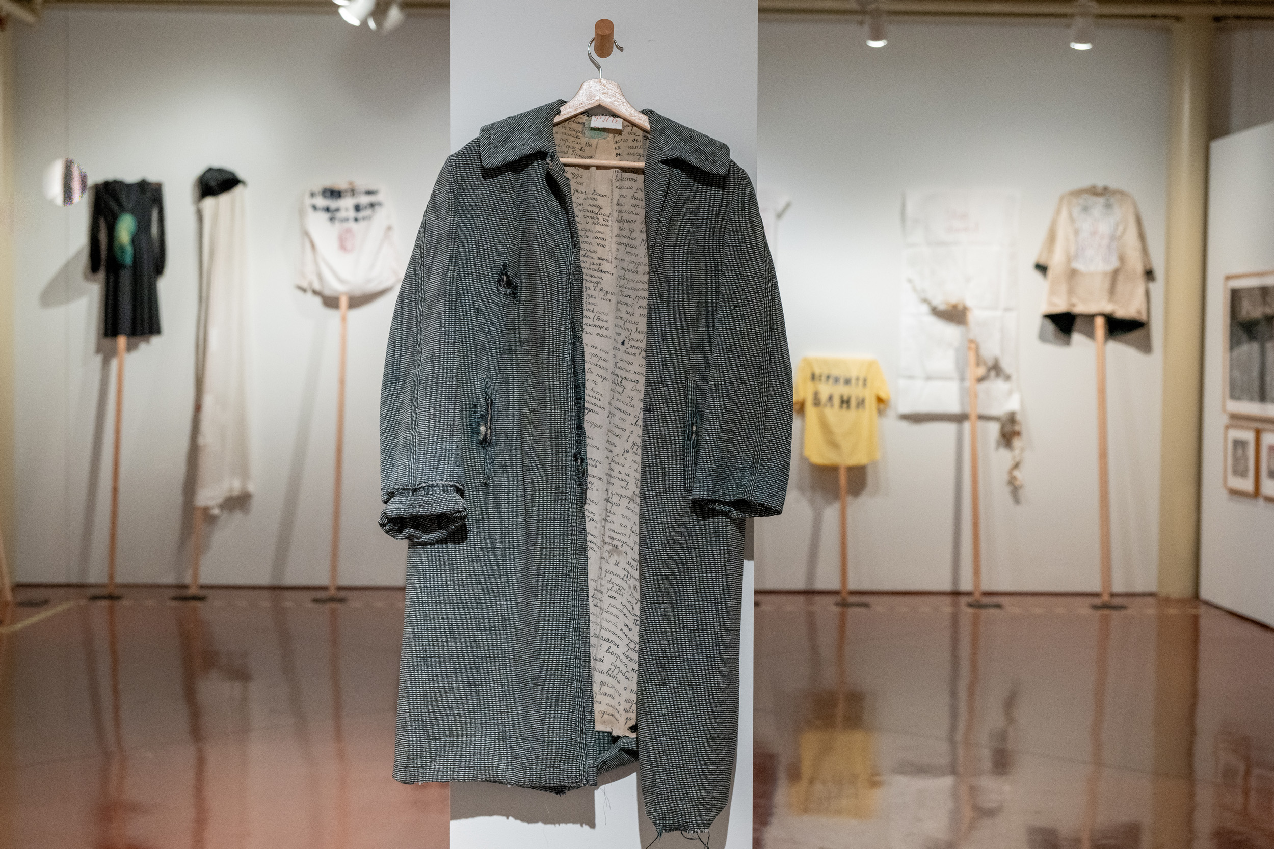 Photograph of a textile art exhibit featuring a gray coat with handwritten text on its inner lining, displayed on a white pillar. Surrounding the coat are various garments and fabric pieces mounted on walls and stands, showcasing diverse textures, colors, and artistic designs in a gallery setting.