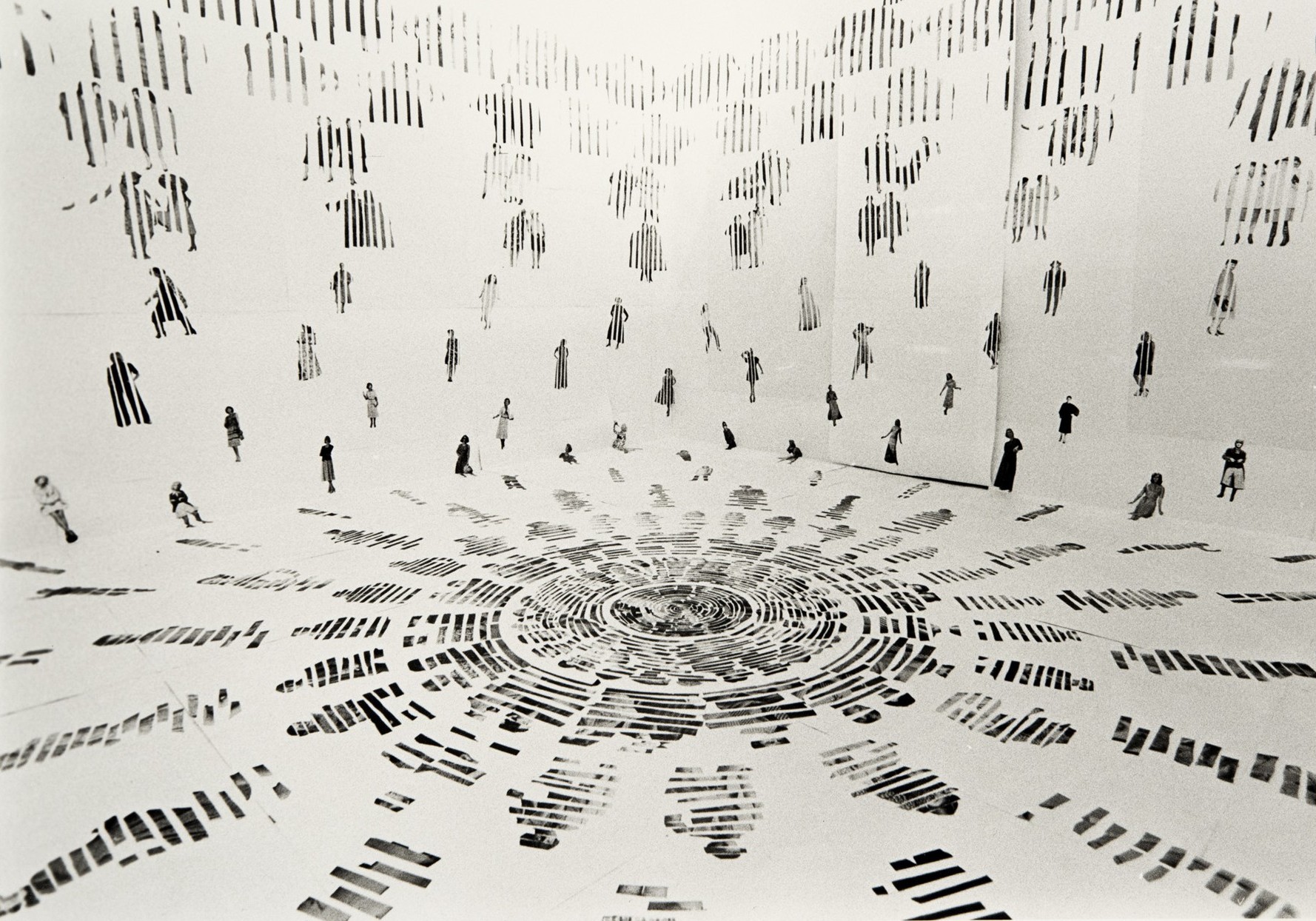 A black and white photograph showing numerous small human figures arranged in a circular pattern on a flat surface, with additional figures suspended vertically around the perimeter. The composition creates a sense of depth and movement, highlighting contrast between light and shadow with striped patterns on figures and ground.