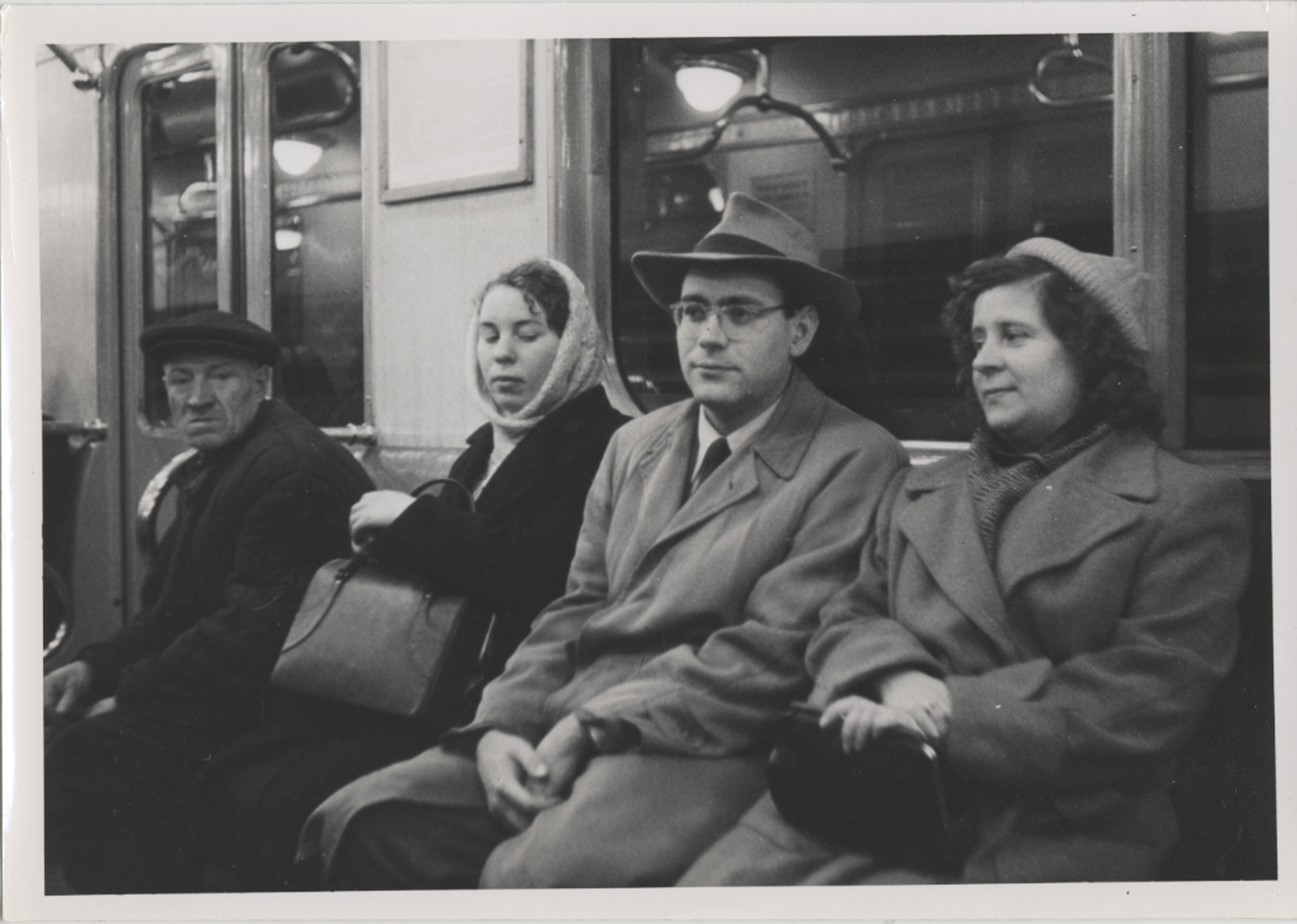 Black and white photograph showing four people sitting closely on a subway or train bench, dressed in mid-20th century attire including coats, hats, and scarves. The setting features reflective windows and overhead lighting, capturing a moment of quiet travel or commute.